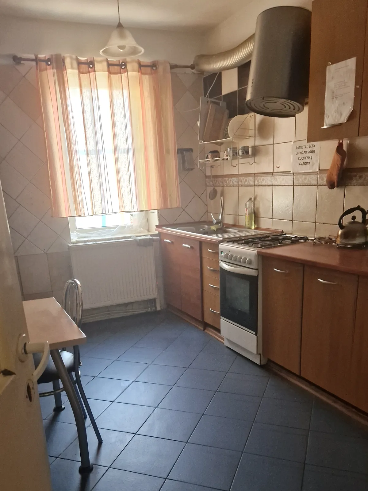 Wspólna kuchnia: kuchenka gazowa z piekarnikiem, blat roboczy, szafki, stolik pod oknem, czajnik.