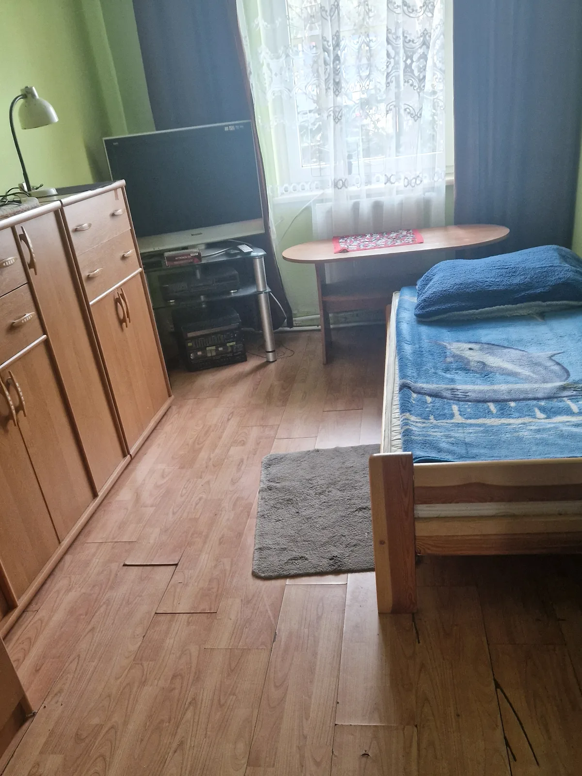 Pokój mieszkalny: łóżko przykryte kocem, telewizor na komodzie, stolik przy oknie, dywanik na podłodze.